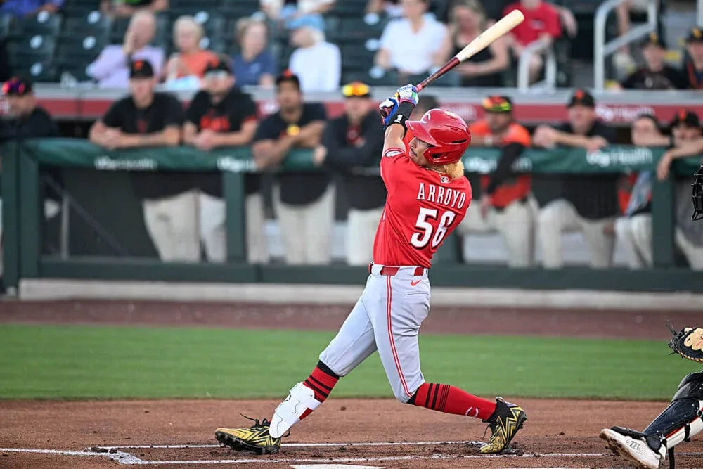 Cincinnati prospect Edwin Arroyo, clad in a red jersey and batting helmet and gray pants, holds his bat high over his head with both hands as he follows through on a swing.
